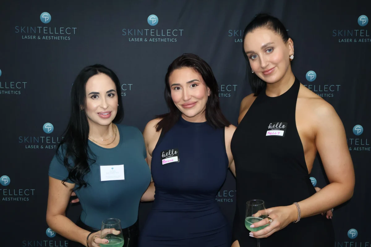 Three women from Skintellect Laser & Aesthetics smiling and posing at a client appreciation event, standing in front of a branded backdrop and holding drinks.